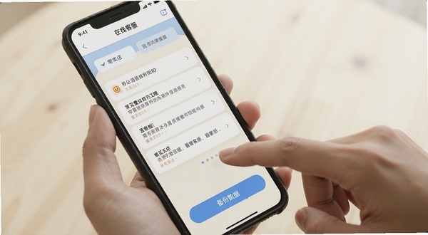秒店应用内的帮助与反馈页面,手机屏幕显示常见问题列表和在线客服入口,背景为柔和的米白色,画面中有一只手正点击屏幕上的“备份数据”按钮,风格写实,光线自然