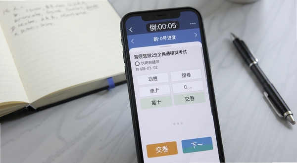 驾照全典通模拟考试界面特写,画面主体为手机屏幕中的考试答题页面,上方显示倒计时与题号进度,中间为单选题题干与选项按钮,底部为交卷与下一题操作栏,背景为柔和浅灰色学习桌面,旁边放置笔记本与签字笔,体现备考场景