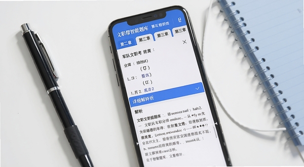 文职帮智能题库页面展示,手机屏幕中显示一道军队文职考试选择题,下方展开详细的文字解析区域,屏幕上方有章节分类标签,背景为干净的白色学习桌面,桌面角落放置一支笔和一本笔记本,整体色调以蓝白为主,突出学习氛围