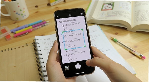 喵喵错题拍照录题功能展示,画面中一部手机正在拍摄数学试卷,屏幕显示识别框自动框选错题区域,背景为书桌场景,有文具和课本点缀,风格写实明亮