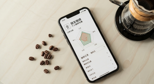 朋友咖啡个人咖啡档案页面,手机屏幕展示口味雷达图与咖啡豆记录列表,背景为淡雅米色桌面,散落几颗咖啡豆与手冲壶,风格简约文艺