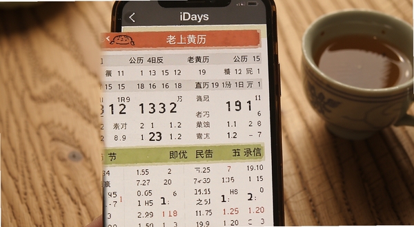 老黄历iDays软件界面截图,展示公历农历双历显示与节气标注,手机屏幕特写,背景为木质桌面与茶杯,温馨生活场景