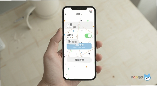 占星猫移动端使用操作示意图,画面展示一只手手持手机,屏幕内为占星猫设置页面,包含通知开关与缓存清理按钮,背景为简洁浅色桌面环境,光线柔和自然,适配Boogu模型生成