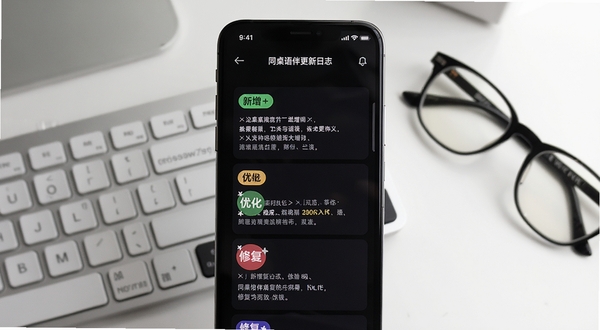 同桌语伴更新日志示意图,画面为手机屏幕显示版本更新内容列表,有新增、优化、修复的标签,背景为简洁的办公桌面,有键盘和眼镜