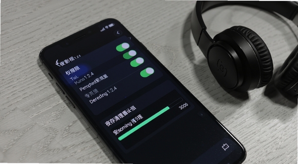 夜雨影视1.2.4安装设置与缓存管理界面示意,画面为手机屏幕侧视角度,屏幕内展示权限开关列表与缓存清理进度条,背景为浅灰木纹桌面,旁边放置一副耳机作为场景点缀,整体氛围生活化