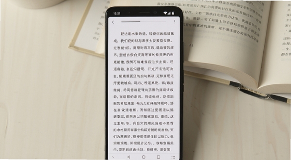 轻阅读软件主界面展示,画面中央是一部竖屏安卓手机,屏幕显示简洁的电子书阅读页面,白色背景配深灰色文字,顶部有细窄的进度条,背景为浅米色桌面与一本翻开的纸质书,整体风格清爽极简,光线柔和,构图居中