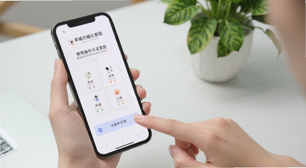 羊城巾帼云家园使用操作示意图,画面主体为手持手机进行点击操作的近景,屏幕显示功能引导页面,背景为简洁的浅灰色桌面与绿植点缀,风格干净明亮,突出上手简单的视觉感受