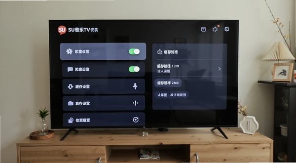 SU音乐TV安装设置界面,电视屏幕上显示权限开关与缓存路径选择面板,UI为深色卡片风格,画面主体为设置列表区域,背景为客厅电视柜场景