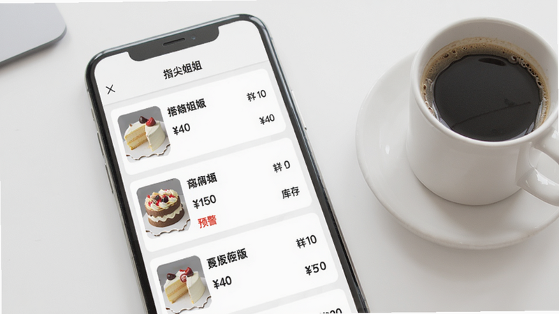 手机界面展示商品管理页面,列表显示多个商品条目,每个商品有缩略图、名称、价格和库存数字,其中一款蛋糕库存显示“预警”红色字体;背景为简约白色桌面,旁边放着一杯咖啡。整体干净、专业,体现指尖姐姐商家版智能库存功能。