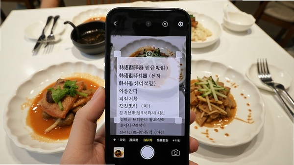 手机拍摄一份韩文菜单,屏幕自动框选文字区域并显示中文翻译,桌面上摆放着韩式料理,画面清晰明亮,强调“韩语翻译器”的拍照识别能力。