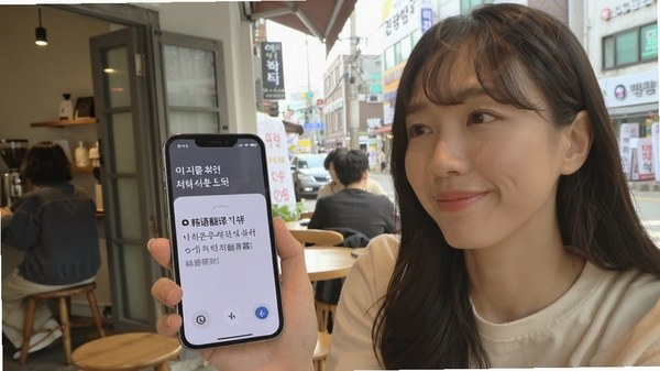 用户对着手机说话,屏幕即时显示韩文翻译和中文释义,背景是首尔街头咖啡馆,人物表情轻松,画面明亮,突出“韩语翻译器”的语音互译功能。