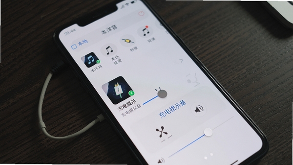 光氪充电提示音自定义编辑界面,手机屏幕显示本地音乐选取列表,一首歌曲正在被拖动到“充电提示音”设置区域,界面有简单的剪切和音量调节滑块,背景是深色木纹桌面,突显个性化操作。
