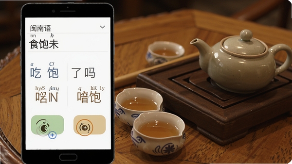 闽南语翻译软件的双向翻译功能演示截图,界面显示闽南语输入“食饱未”和普通话翻译结果“吃饱了吗”,并列出拼音标注和发音按钮,背景为闽南茶桌场景,茶杯与茶壶点缀,画面清晰,突出翻译准确性与学习辅助功能。