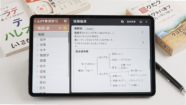 饭团子日语JLPT单词学习界面展示,左侧是单词列表,右侧是详细释义和语法树状图,背景为简洁的白色书桌,散落着几本日语教材和一支笔,风格写实且专业,突出备考学习氛围