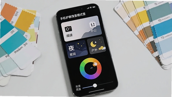 手机护眼情景模式设置页面,屏幕显示四种模式卡片(阅读、办公、夜间、户外),每种卡片图标不同,下方有色温调节色环和亮度滑条,背景为浅灰色桌面,手机旁边摆放着不同颜色的色卡,画面明亮清晰,突出个性化调节功能