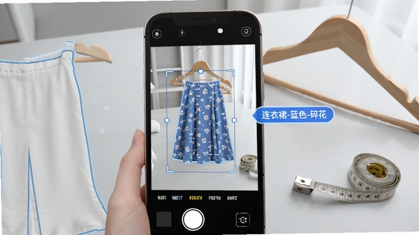 z衣橱智能识别功能预览,手机屏幕显示拍摄裙子的界面,画面中识别框捕捉衣物轮廓,右侧弹出自动分类标签如“连衣裙-蓝色-碎花”,背景为简洁明亮的浅灰色桌面,旁边放有衣架与卷尺,突出科技感与便捷性。