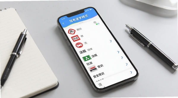 驾考速学精灵软件在手机上展示题库列表,题目分类清晰,包含交通标志、法规、安全常识等,背景为浅灰色桌面,旁边放有笔记本与笔,风格明亮写实,突出刷题场景。