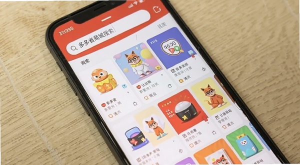 手机屏幕显示多多省商城搜索商品界面,顶部搜索框有文字,下方网格状商品卡片,色彩丰富,模拟真实使用场景,背景为浅色木纹桌面