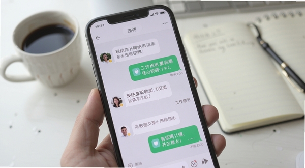 现结兼职软件聊天沟通界面,画面主体为手机屏幕特写,屏幕中显示着用户与招聘方关于工作细节的对话气泡,背景区域为简洁的办公桌面,桌面上放着一杯咖啡和一本记事本,整体氛围轻松高效