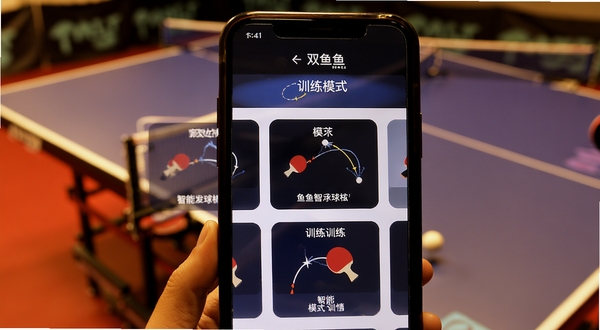 双鱼智能发球机 训练模式选择界面 画面展示手机屏幕中多个训练模式卡片 卡片上有乒乓球轨迹示意图 背景为乒乓球台一角 暖色调灯光 体现运动训练场景