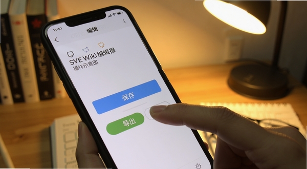 SVE Wiki 编辑器 操作示意图 手指点击手机屏幕上的词条编辑按钮 界面显示保存与导出选项 背景为书桌场景 手机斜放 暖色台灯光线 突出操作细节