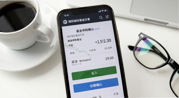 国投瑞银基金交易页面特写,画面中手机屏幕显示基金申购确认页面,上方有基金名称和净值数字,下方有买入按钮和份额确认信息,背景为干净的白色办公桌面,旁边放着一杯咖啡和一副眼镜,营造专业理财场景