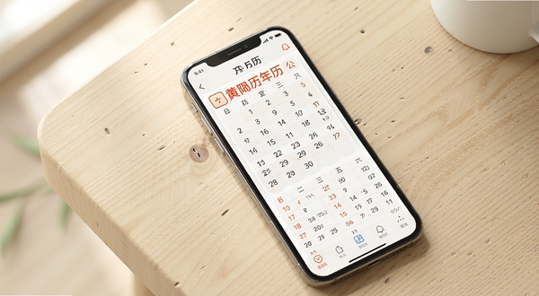 极佳万年历应用主界面展示,画面中央为手机屏幕内显示公历农历对照与黄历宜忌信息,背景为浅色木质桌面,柔和自然光从左上角照入,整体风格简洁清新,突出日历应用核心功能