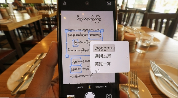 缅甸语翻译通拍照翻译操作示意图,手机摄像头对准一份缅甸语菜单,屏幕中菜单文字被识别框选中并显示中文翻译浮层,背景为餐厅桌面环境,画面色调温暖自然