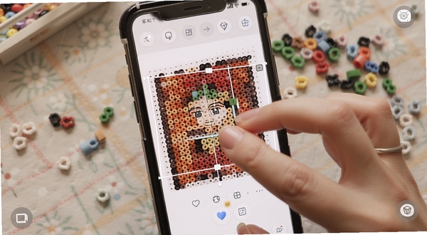 拼豆钻石画触控缩放与网格预览操作界面,画面主体为手指在手机屏幕上双指缩放拼豆图纸,背景区域为柔和的手工桌布和几颗散落的拼豆珠子,细节清晰