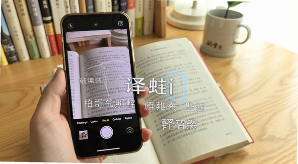 译蛙ai 拍照翻译功能演示 手机摄像头对准一本外文书籍 屏幕上叠加显示中文翻译结果 背景为书桌场景 光线明亮 突出AR翻译效果