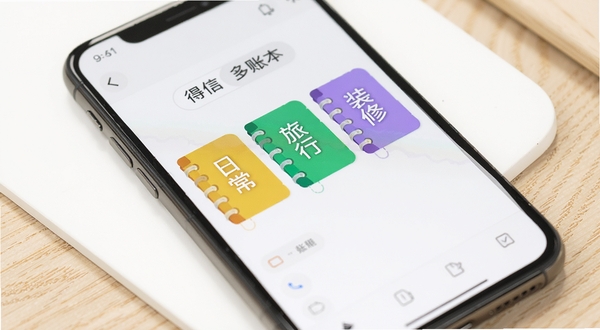 得信记多账本管理界面特写,画面主体为手机屏幕中并排显示三个彩色账本标签,标签上分别标注日常、旅行、装修字样,背景区域为纯白色桌面与浅木色纹理,光线柔和,突出账本切换的便捷感