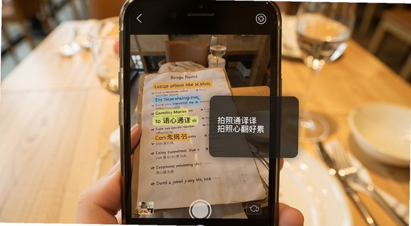 语心通译拍照翻译场景示意图,画面中心为手机摄像头取景框对准英文菜单,取景框内文字被高亮识别并显示中文译文浮层,背景区域为餐厅桌面环境虚化,整体色调温暖明亮,体现实用性与便捷性,适配Boogu模型生成。