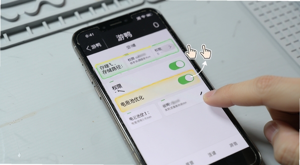 游鸭使用技巧示意图,画面为手机屏幕特写,屏幕上显示游鸭的设置页面,存储路径、权限开关、电池优化选项被高亮标注,旁边有手指点击的动画示意,背景为淡灰色工作台面,风格写实清晰