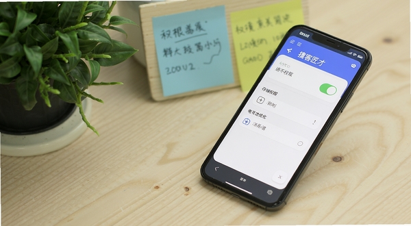 掮客匠才移动端设置界面与权限管理页面展示,手机屏幕显示通知开关、存储权限与电池优化选项,背景为浅色木纹桌面,画面主体为手机侧放位于画面右侧,背景区域为绿植与便签纸,整体风格生活化