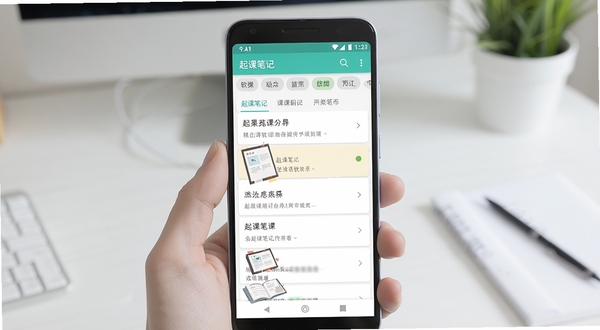 起课笔记 软件主界面功能全貌展示 画面主体为一部居中放置的安卓手机 背景区域为简洁的书桌场景 手机屏幕上显示起课笔记的笔记列表与分类标签 画面风格清新明亮 适配Boogu模型生成