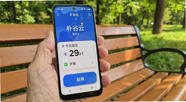 一部安卓手机展示朴谷云应用首页,大号字体显示今日血压和步数,界面按钮宽大醒目,手机放在老人手中,背景是公园长椅和绿树,阳光明媚,突出适老化设计。
