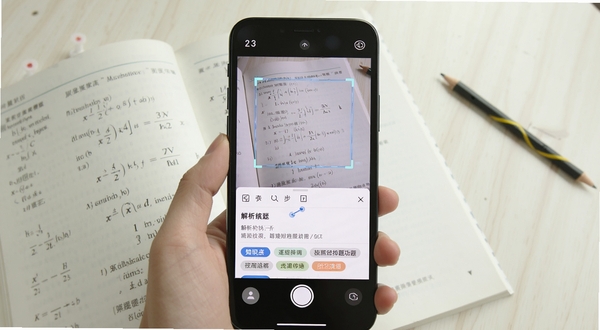 题库搜题拍照搜题功能演示,手机屏幕显示正在识别一本打开的数学练习册,识别框精准框选题目,屏幕下方弹出解析结果和知识点标签,画面明亮清晰,突出智能识别过程。