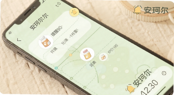 安珂尔用药提醒功能界面,手机屏幕显示时间轴与药品图标,有铃铛提醒标志,背景为淡绿色,画面温馨,带有安珂尔品牌字样