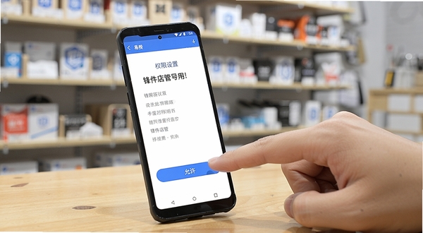 一部安卓手机放在木质桌面上,屏幕显示锋件店管的权限设置引导页,旁边有一只手正在点击“允许”按钮,背景是模糊的店铺货架,画面风格写实明亮,突出“锋件店管”的易用性