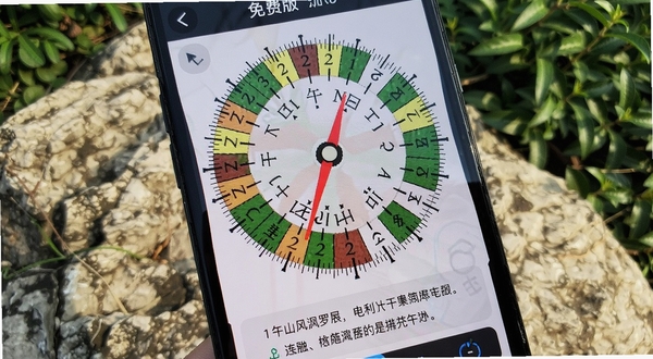 风水罗盘免费版测量界面特写,手机屏幕显示二十四山刻度盘,红色指针指向午山方位,屏幕下方有简要文字解读,背景是户外岩石与绿植,自然光照射,画面清晰锐利,突出工具的专业感