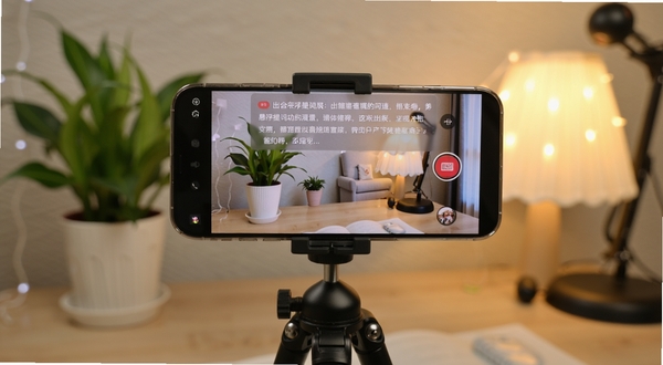 出镜助手悬浮提词功能展示,画面主体是一部手机架在三脚架上,手机屏幕显示正在录制视频,屏幕上方悬浮着半透明的文字提词框,背景是温馨的室内直播场景,有绿植和柔和的灯光,细节清晰,风格写实,适配Boogu模型生成