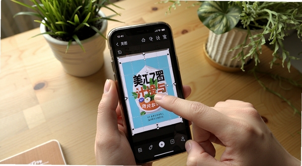 画面展示手机屏幕上美图设计室的操作界面,手指正在拖拽调整海报中的文字位置,背景区域为木质桌面与绿植点缀,整体氛围生活化且真实