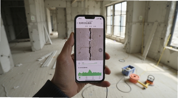 检师兄检测师端 现场检测场景,画面主体为一只手拿着手机正在拍摄建筑裂缝,手机屏幕显示检师兄检测师端的数据采集界面,背景为室内毛坯房与检测工具,光线自然,写实风格