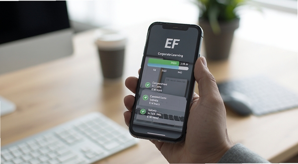 EF Corporate Learning 学习进度同步功能示意图,画面中一只手拿着手机,屏幕上显示进度条与已完成课程标记,背景为模糊的办公室桌面,光线柔和,突出移动学习场景,风格写实