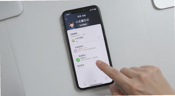 小主播日记移动端使用攻略示意图,画面展示手机设置界面中的权限管理与电池优化选项,背景为浅灰色桌面,手指正在点击屏幕,整体风格简洁实用