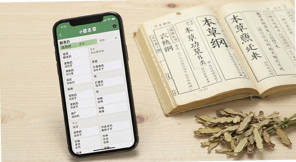 e德本草软件内部的中药材列表界面,手机屏幕显示着按笔画或功效分类的药材目录,如“解表药”、“清热药”等,画面清晰明亮,背景为浅色木纹桌面,旁边放着一本翻开的纸质本草纲目书籍和一株干燥的草药,体现传统与现代查询方式的对比。