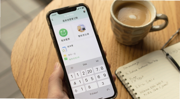 易来钱管家记账界面特写,手机屏幕显示分类图标与数字键盘,背景为温暖木质桌面,旁边放着一杯咖啡和一本笔记本,营造日常记账场景