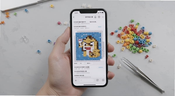 天天拼豆新手操作示意图,画面中一只手拿着手机,屏幕上显示拼豆图纸详情页,旁边有彩色拼豆颗粒和镊子作为点缀,背景为浅灰色桌面,整体风格写实清晰