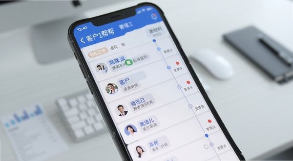 企管帮客户管理模块界面特写,画面主体为手机屏幕居中显示客户列表与跟进时间轴,背景区域为淡灰色办公桌面虚化,屏幕内突出商机阶段标签与提醒红点,整体色调以蓝白为主,细节清晰,体现移动办公场景