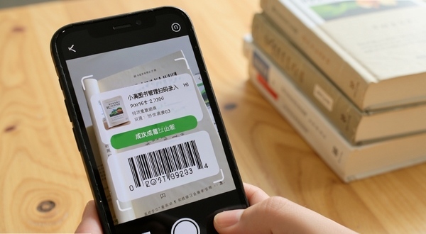 小满图书管理扫码录入功能特写,画面主体为手机摄像头对准书籍条形码,屏幕显示识别成功的书籍信息卡片,背景为木质桌面与几本叠放的书籍,光线明亮柔和